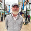 Man in sweater and ball cap with white beard stands on busy street
