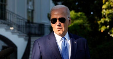 Joe Biden stands with dark sunglasses in a blue suit in front of the White House