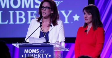 Tiffany Justice in a blue dress and white blazer speaks with Tina Descovich in a red dress in front of a Moms for Liberty logo