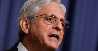 Merrick Garland with white hair in a suit and wearing glasses stands in front of a blue background