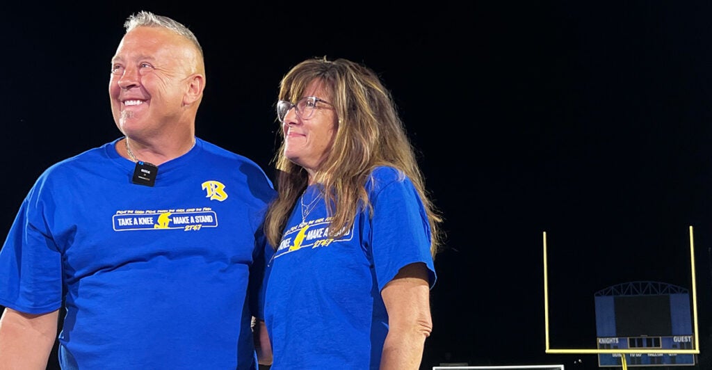 Coach Joe Kennedy stand on a football field with his wife by his side.