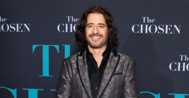 Actor Jonathan Roumie smiles on the red carpet in a grey jacket and a black shirt.