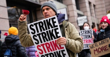 Man in jacket holds sign reading "Strike" while shaking a cowbell.