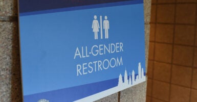 A dark and light blue bathroom sign says "ALL-GENDER RESTROOM" with two white man and woman images, side-by-side.