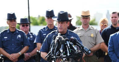 A sheriff speaks at a press conference with multiple microphones surrounded by other officers in uniform.