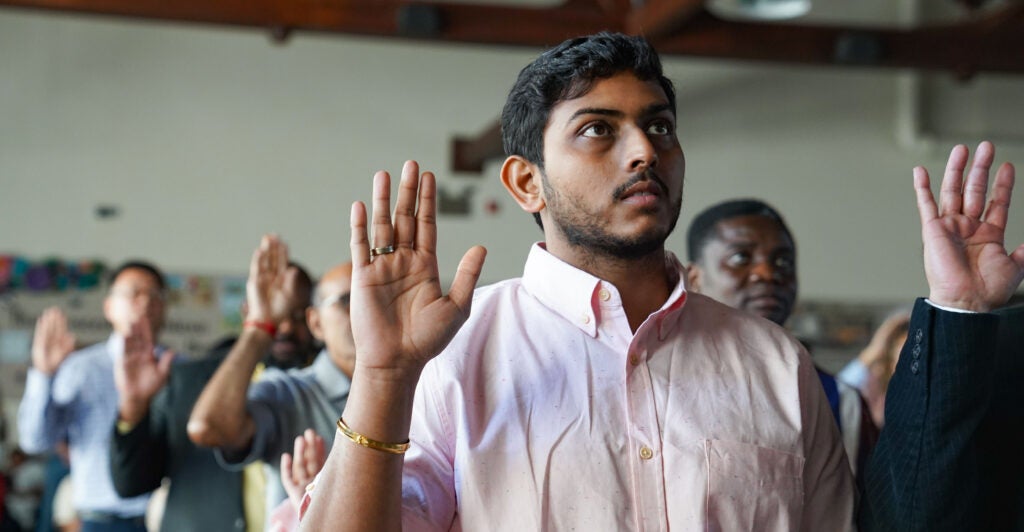 A young man with black hair and brown skin wears a light pink shirt as he raises his right hand with a gold ring on it during his naturalization ceremony.