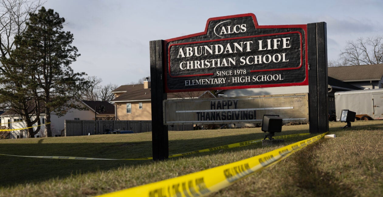 Yellow crime scene tape drapes around a black school sign with white lettering for Abundant Life Christian School.