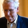 Mitch McConnell, in a blue suit, walks down an orange Senate hallway, with a frown on his face.