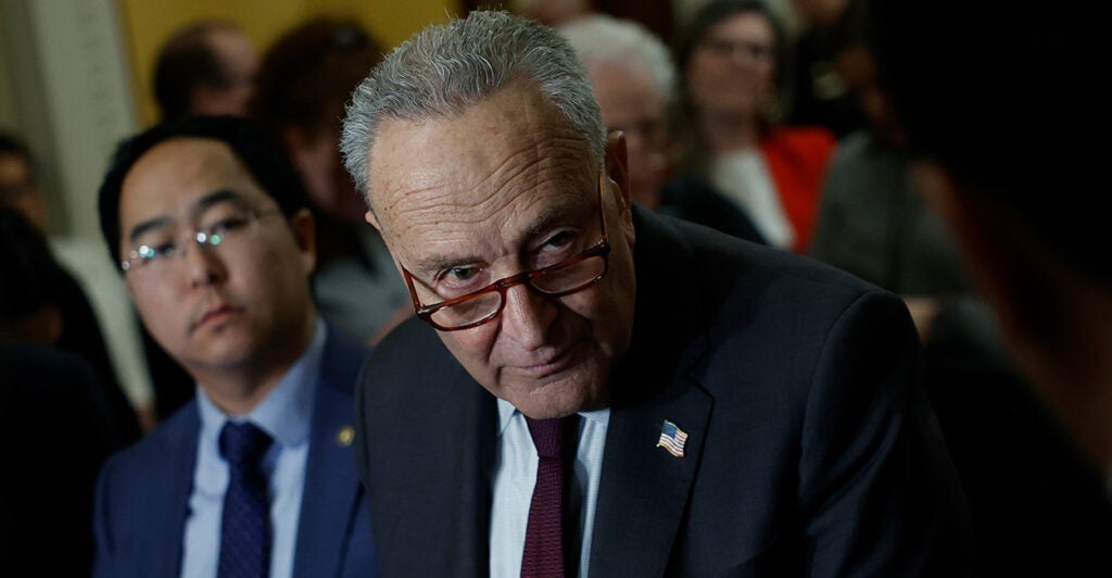 Senate Majority Leader Chuck Schumer, in a black suit and glasses, stares ominously during a conference.