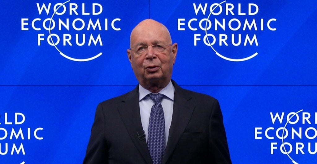 Klaus Schwab stands in front of a World Economic Forum banner.