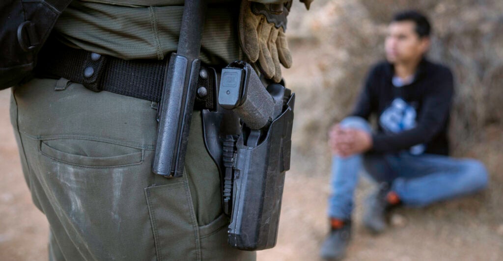 A U.S. Border Patrol agent, dressed in a green uniform with a black holstered gun, stands near a detainee, sitting in a black shirt.