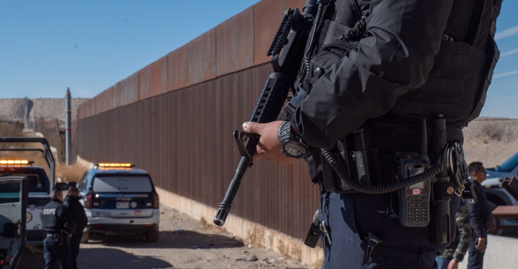 An officer dressed in a black uniform holds a gun pointed down.