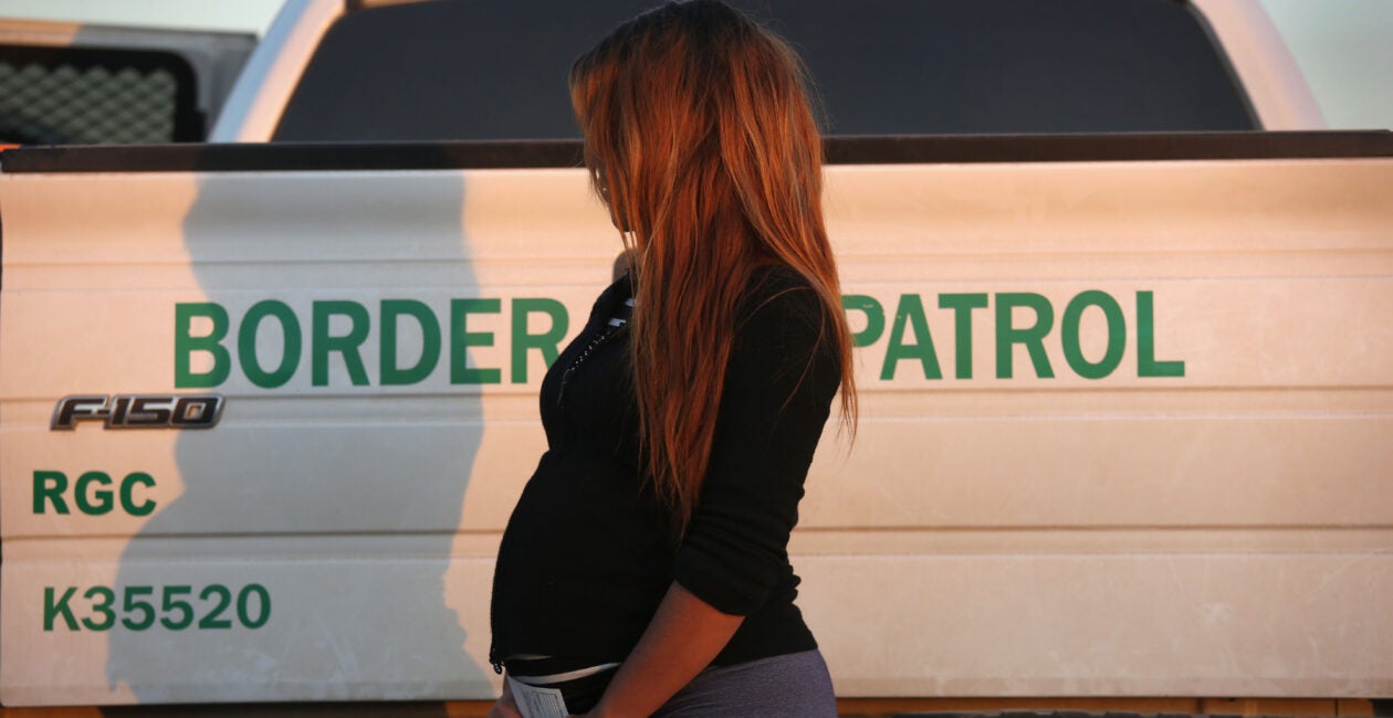 A pregnant women stands in a black shirt next to a white U.S. border patrol truck.
