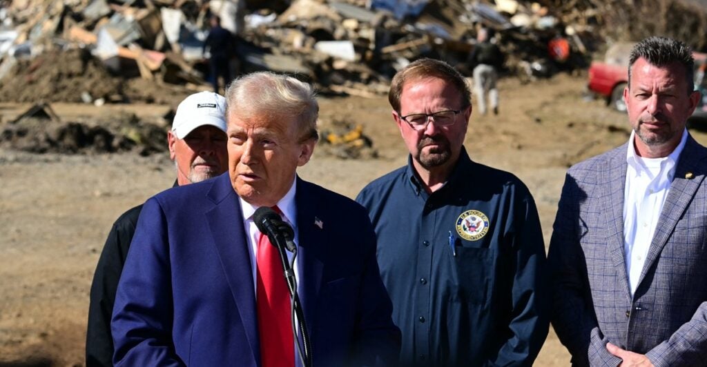 Donald Trump stands in the middle of a hurricane devastated area in a suit and tie.