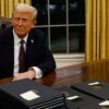 Donald Trump sits at his desk in a dark suit with stacks of executive orders.