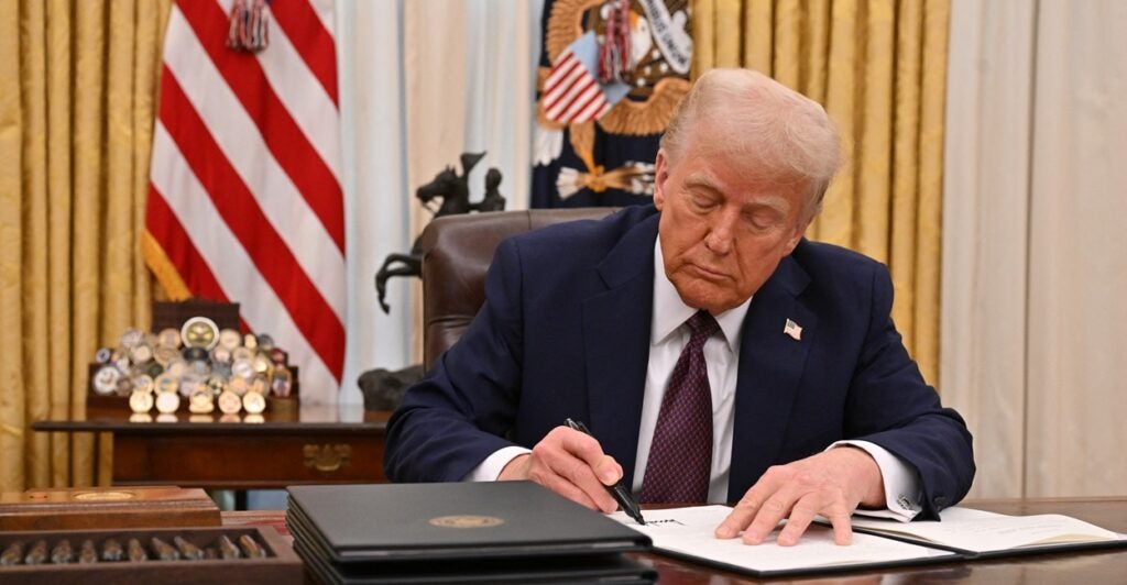 Donald Trump sits at his desk and signs a piece of paper while staring down at it.