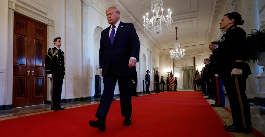 Donald Trump walks with his hands to his side in the White House on a red carpet.