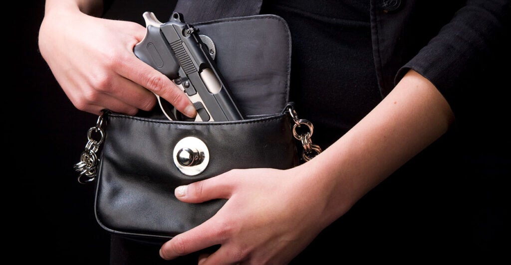 A woman's hands reach into a black purse to pull out a black concealed handgun