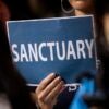 A woman holds up a sign that says "SANCTUARY" at a meeting.