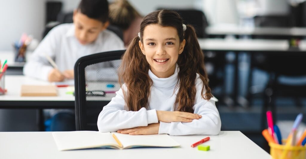 A young girl with a ponytail sits at a desk with her hands folded in front of her.