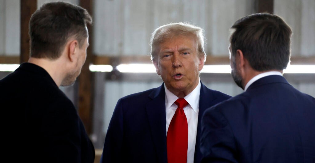Donald Trump in a suit speaks with J.D. Vance and Elon Musk
