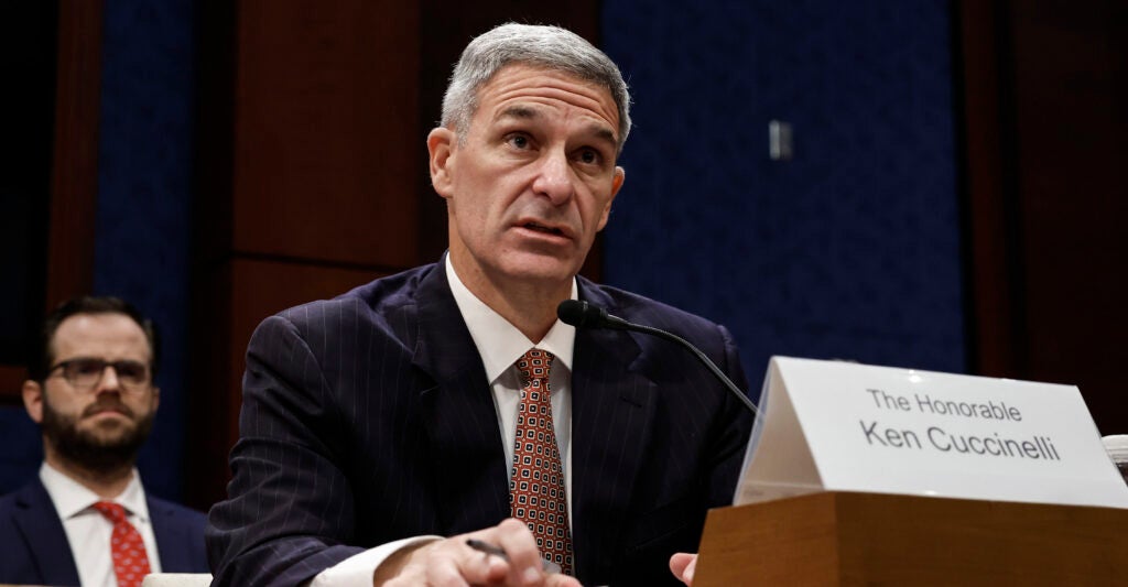 Ken Cuccinelli in a dark suit and sitting at a table testifying to a congressional committee