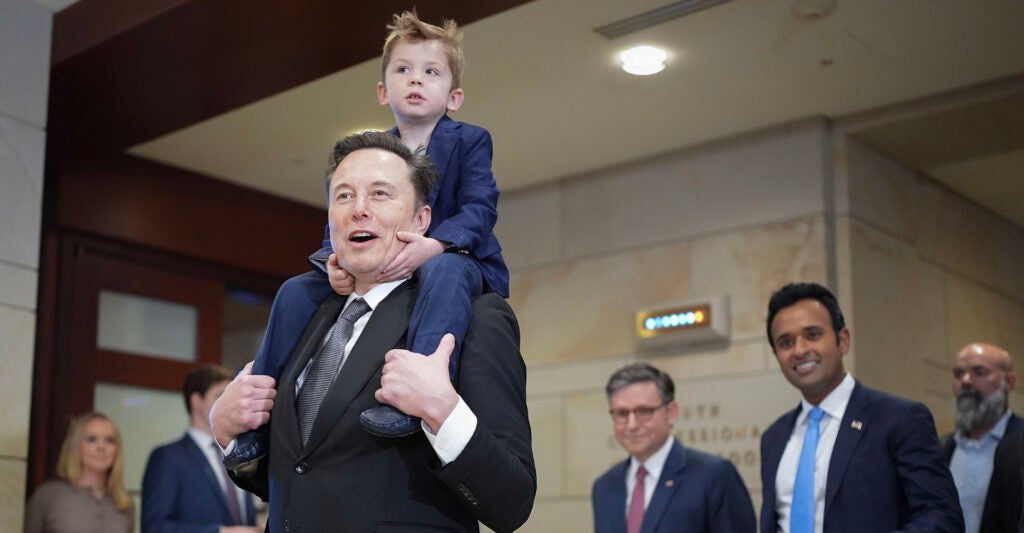 Elon Musk, dressed in a black suit, carries his young son in a blue suit on his shoulders.