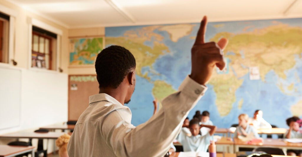 A black male teacher stands in front of a classroom wearing a white dress shirt.
