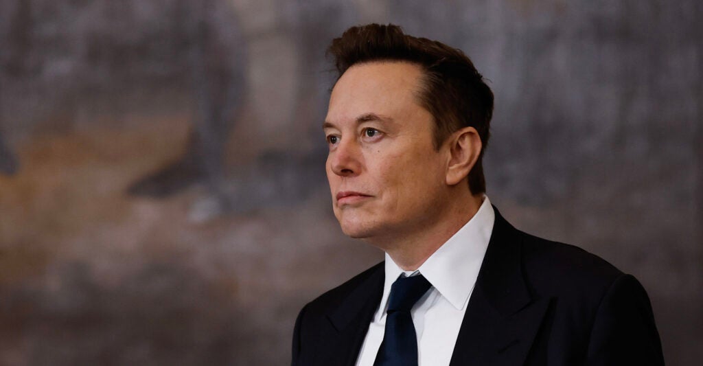 Elon Musk looks on, wearing a black suit, black necktie, and a white dress shirt.