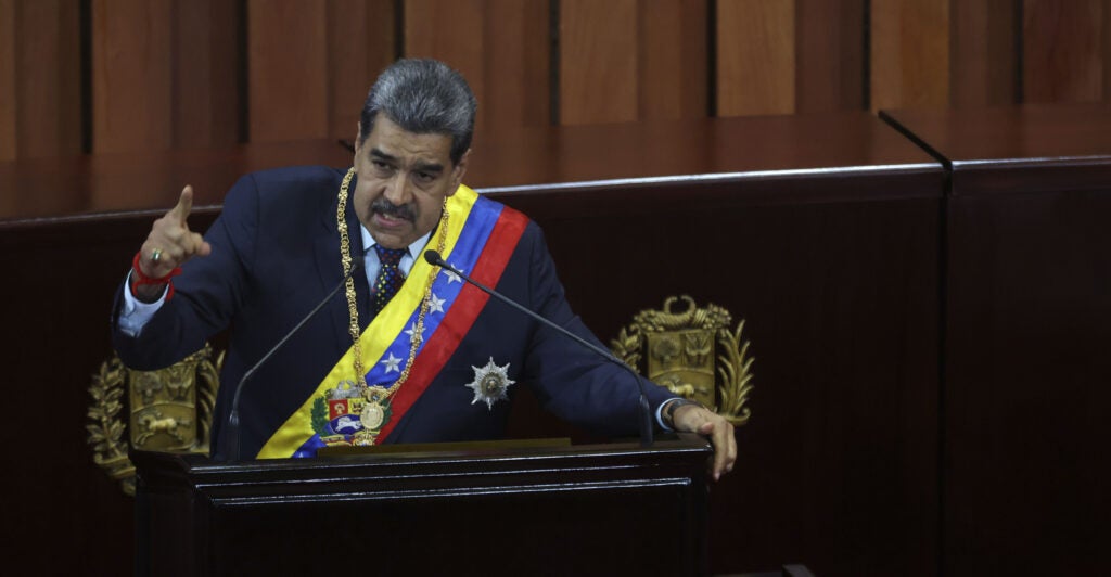 Nicolás Maduro stands in navy blue official attire including a sash across his body decorated in the colors of the Venezuelan flag.