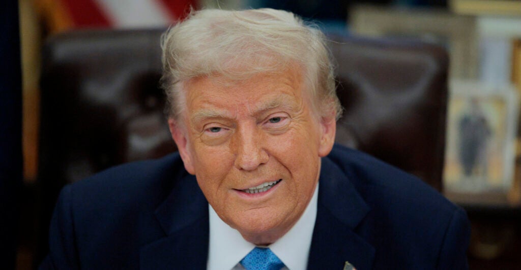 Donald Trump smiles, wearing a dark blue suit and light blue necktie.