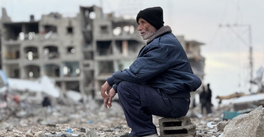 A Palestinian man sits on a gray cinderblock, wearing a black winter hat, blue winter coat, and blue pants.