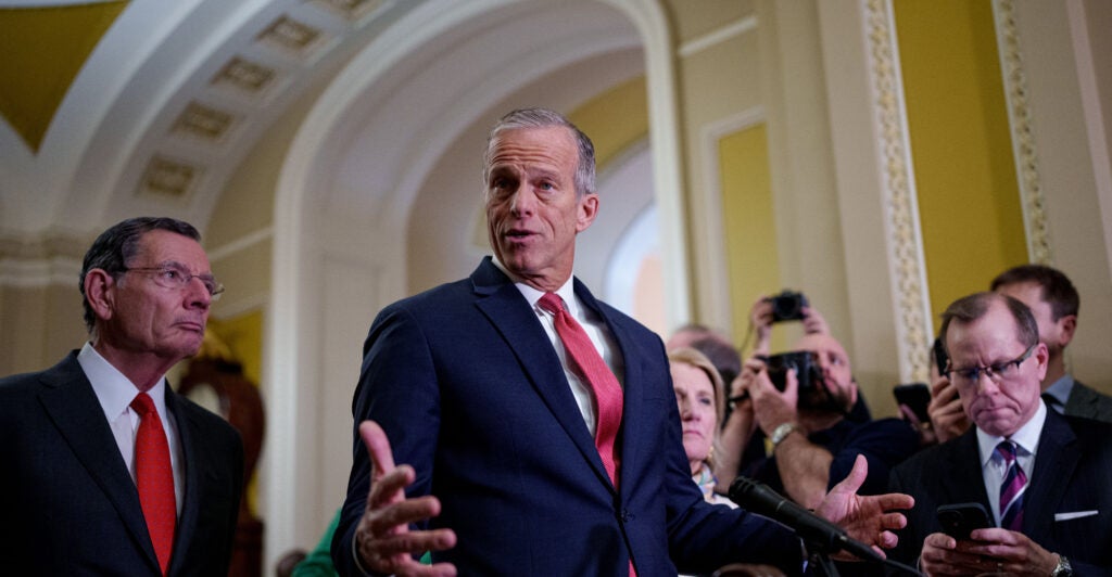 John Thune gestures, wearing a dark blue suit and red necktie.