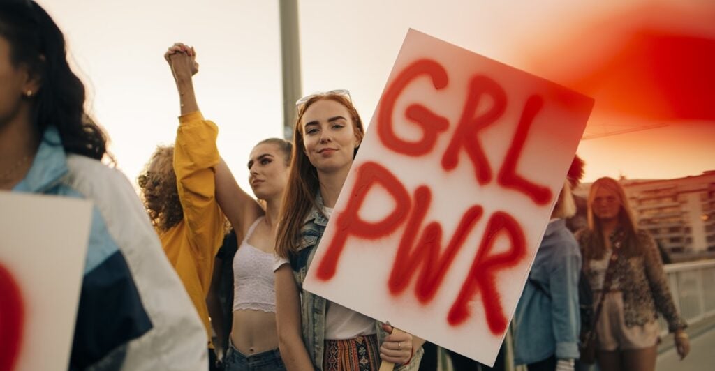 A college-aged woman holds a sign that says, "GRL PWR".