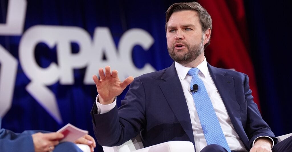 JD Vance raises his right hand as he sits and talks during an interview.