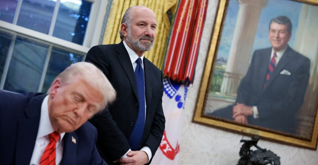 Howard Lutnick stands with his hands folder as Donald Trump signs an executive order.