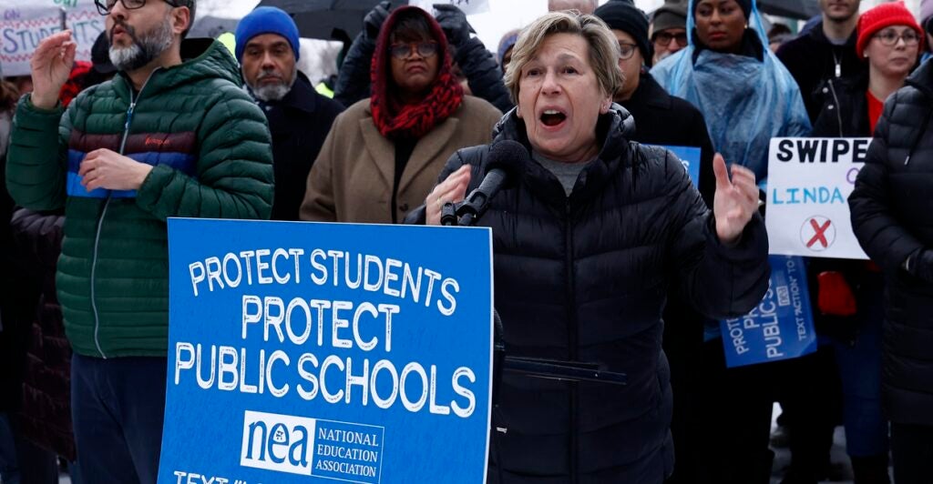Randi Weingarten is standing outside wearing a winter jacket as she shouts during a protest near a sign.