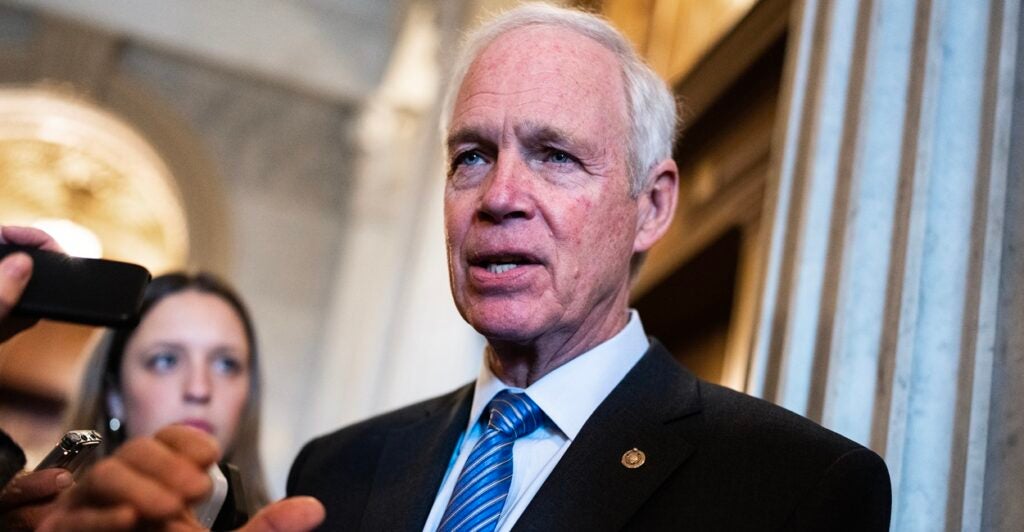 Ron Johnson stands in a room and looks to his left while surrounded by reporters.