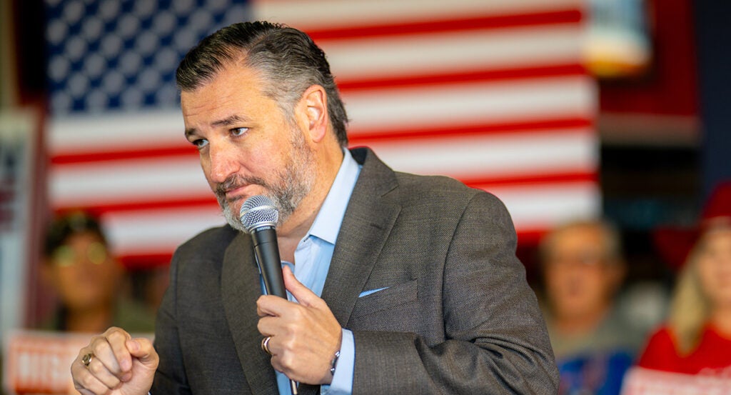 Ted Cruz in a suit without a tie leans in in front of an American flag