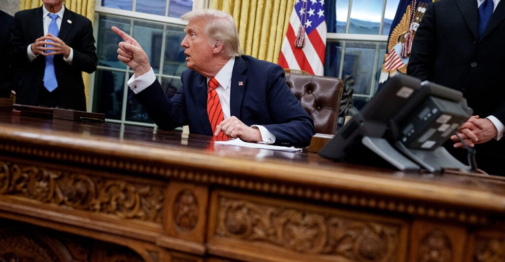 President Donald Trump, seated behind his desk in the Oval Office, pointing at someone