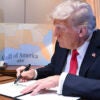 Donald Trump in a blue suit with a red tie signs an executive order with a map reading "Gulf of America" in the background.