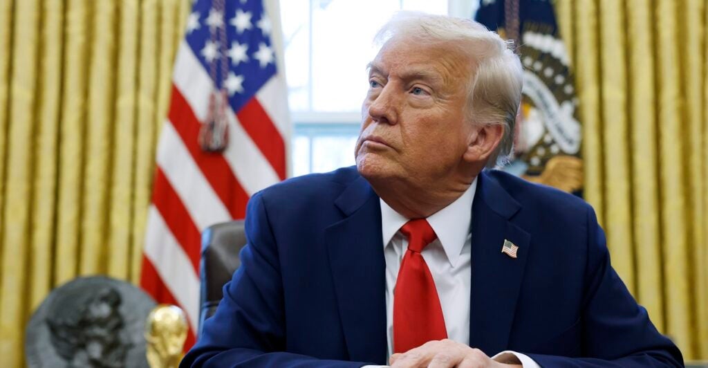Donald Trump sits at a table with his hands set in front of him while he looks to his right.