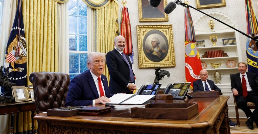 With microphones around him, Donald Trump sits at his desk and talks to reporters.