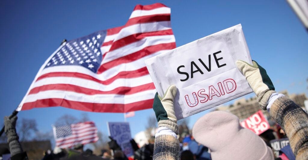 A pair of hands hold up a sign that states "SAVE USAID" as an American flag waves in the background.