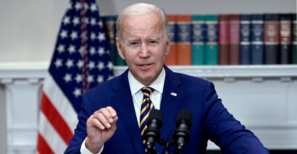 Joe Biden in a blue suit speaking at a podium with the American flag and books on a shelf behind him