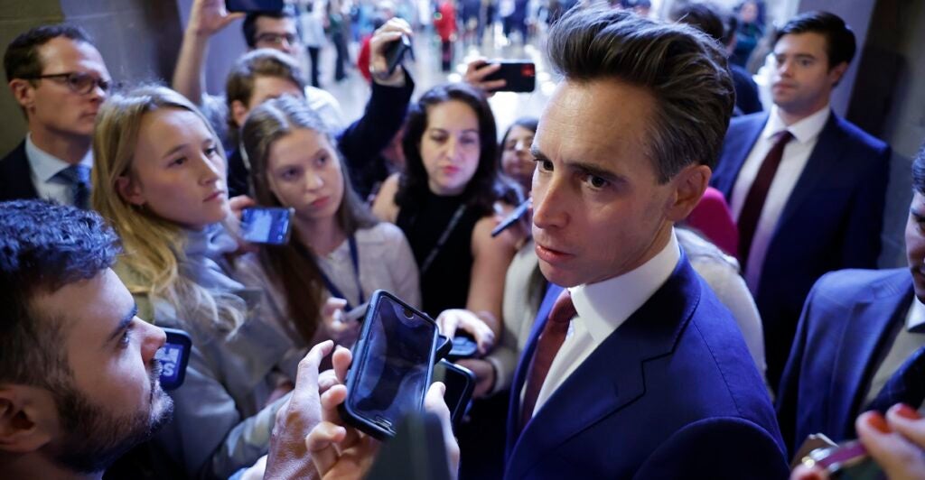 Josh Hawley is standing outdoors surrounded by reporters putting their microphones in front of his face.