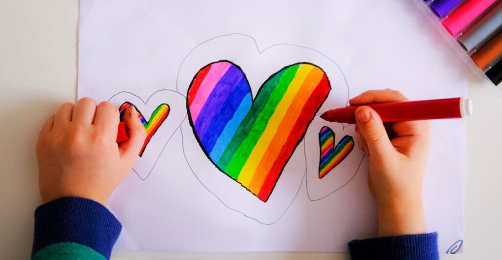 a child's hands holding a marker coloring in hearts with the LGBTQ rainbow