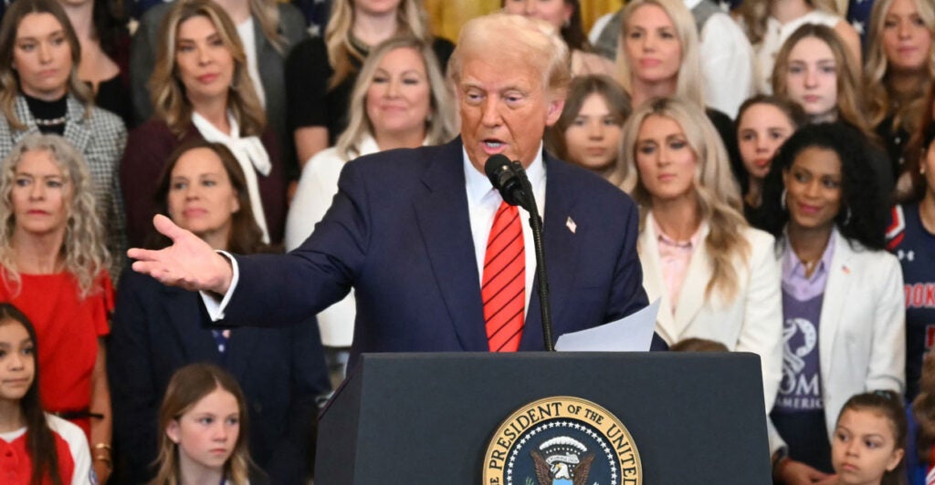 Donald Trump in a blue suit and red tie at the podium gesturing toward the crowd with several women standing behind him