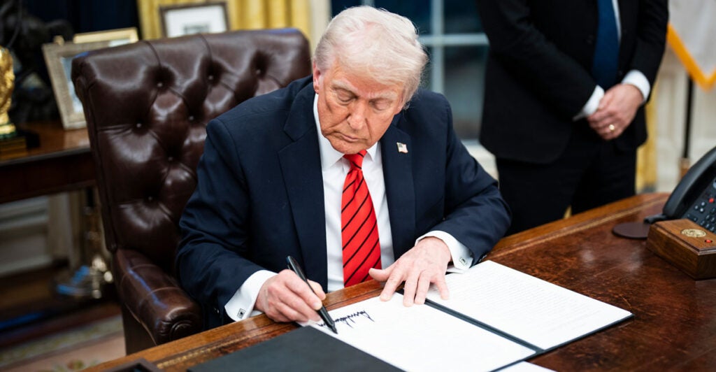 Donald Trump in a blue suit and red tie at the oval office desk signing an executive order