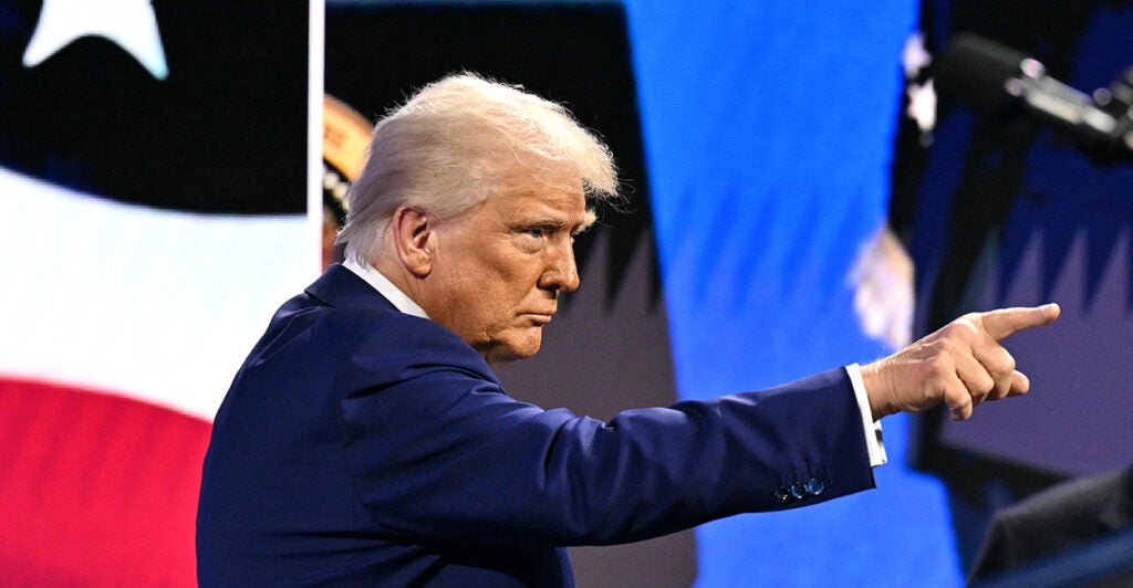 Donald Trump in a dark suit looking out and pointing to the crowd, with a large American flag in the background